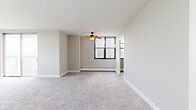 This single-floor residence offers a thoughtfully arranged layout with multiple living and sleeping areas, all finished with neutral-toned carpeting and crisp white walls that lend a clean, airy feel throughout. The spacious living area is anchored by a ceiling fan with an integrated light fixture and receives natural light through a large multi-pane window. Sliding glass doors open to a balcony, framing sweeping outdoor views that include treetops and a body of water in the distance — a striking visual feature visible from multiple rooms.

The bedroom areas are generously proportioned, each benefiting from large windows equipped with blinds that showcase the same scenic water and greenery views. A wall-mounted air conditioning unit and baseboard heating system ensure year-round climate comfort. Closet space is provided with a built-in rod and shelf system behind louvered doors.

The kitchen is equipped with warm wood cabinetry, granite-style laminate countertops, a dishwasher, electric range, and a double sink set on white tile flooring. The bathroom features white tile flooring, a tub-shower combination with white subway tile surround, and a vanity with cabinet storage below.