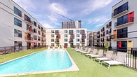This outdoor amenity space in Houston, TX offers a well-appointed environment designed for both relaxation and social gathering. At its centerpiece is a generously sized rectangular swimming pool with clear, blue water, framed by a clean concrete pool deck and surrounded by artificial turf that adds a lush, green aesthetic. Rows of white lounge chairs are arranged poolside, providing ample seating for sunbathing beneath open skies, with views of the surrounding Houston skyline and neighboring high-rise construction.

The space is enclosed within a modern multi-story building complex characterized by a contemporary facade featuring light gray siding, colorful accent panels in red, orange, and yellow, and private balconies with black metal railings. The vibrant pops of color lend the property a distinctive, urban character.

A separate courtyard area features wide concrete pathways intersecting with patches of manicured artificial turf, creating a flexible open-air gathering space. A large, striking mural of a stylized figure adorns one building wall, serving as a prominent artistic focal point. Floor-to-ceiling glass-fronted amenity rooms are accessible from the courtyard, enhancing the connectivity between indoor and outdoor spaces. The overall ambiance is energetic, modern, and community-oriented.