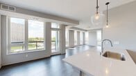 This well-appointed 2-bedroom, 2-bathroom unit offers a thoughtfully designed open-concept layout spread across a single floor. The living room is anchored by expansive floor-to-ceiling windows that flood the space with natural light and frame sweeping urban and city skyline views. Dark hardwood flooring runs throughout the main living areas, adding warmth and contrast against the neutral gray walls and white trim. A balcony door in the living area provides direct outdoor access and additional light. The kitchen is a standout feature, showcasing a large white quartz island with an integrated sink, white cabinetry with a subway tile backsplash, stainless steel appliances including a gas range and French door refrigerator, and stylish glass pendant lighting overhead. The bedrooms are carpeted for comfort, with the primary bedroom benefiting from a large window offering city views. A dedicated closet provides valuable storage. Both bathrooms feature tub-shower combinations and wood-look flooring; one is finished with classic white subway tile surround, while the other offers a clean, minimalist design. Track lighting and pendant fixtures throughout contribute to the unit's modern, urban aesthetic.