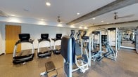 This fitness center offers a thoughtfully designed workout environment that balances functionality with an inviting atmosphere. The space is anchored by exposed wooden ceiling beams that add a warm, architectural contrast to the otherwise clean, contemporary interior finished in neutral tones. Recessed lighting and ceiling fans maintain a comfortable and well-lit environment throughout.

The cardio zone is a standout feature, lined with a row of Life Fitness ellipticals, treadmills, and upright bikes positioned in front of expansive floor-to-ceiling windows. These windows frame a lush outdoor courtyard and deck area, allowing natural light to flood the space and providing an engaging view to keep workouts motivating.

The strength training area is equally well-appointed, featuring a multi-station cable machine and Smith machine, a selection of weight benches, and a full rack of dumbbells. Stability balls are neatly stored nearby, and a large wall mirror enhances the sense of openness while allowing users to monitor their form. A wall-mounted flat-screen TV adds convenience for those who prefer entertainment during their sessions.

The durable rubber-composite flooring throughout provides both safety and comfort underfoot. The overall layout is spacious and thoughtfully organized, offering dedicated zones for cardio, strength, and functional training.