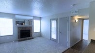 This single-floor residence in Irving, Texas offers a comfortable and functional layout designed for everyday living. The living area serves as the heart of the home, highlighted by a classic brick fireplace that adds warmth and character to the space. Plush wall-to-wall carpeting contributes to a cozy, inviting atmosphere, while a textured popcorn ceiling adds a traditional finishing touch overhead. Natural light filters generously into the room through multiple windows fitted with blinds, creating a bright and airy feel throughout the day. A glass-paneled door provides access to an outdoor area, further enhancing the connection between interior and exterior spaces. The neutral color palette of soft grays and whites keeps the space feeling open and versatile, making it easy to personalize with furnishings and décor. A large mirror above the fireplace mantle visually expands the room, reflecting light and adding depth. The adjoining area, visible from the living room, suggests an open flow into additional living or kitchen space, contributing to the overall functional design of this Irving home.