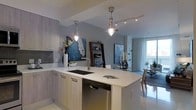 This single-floor space showcases a thoughtfully designed open-concept layout that connects the kitchen, dining, and living areas in a cohesive, contemporary setting. The kitchen serves as a central hub, featuring a large island with a smooth white countertop, an integrated sink, and a stainless steel dishwasher. Light wood cabinetry with brushed metal hardware adds warmth while maintaining a clean, modern aesthetic. A tile backsplash behind the range area introduces subtle texture. Overhead, a combination of industrial-style pendant lights and a track lighting system provides layered illumination throughout the space. The open layout transitions naturally into the dining and living areas, where curated artwork, including a striking blue abstract piece, adds visual depth and personality. A dark wood bookshelf offers display and storage, while candle arrangements on a side table contribute to the ambiance. Sheer curtains near floor-to-ceiling windows allow soft natural light to filter in from what appears to be an outdoor balcony or terrace. The overall atmosphere is contemporary and polished, with a neutral color palette punctuated by carefully chosen decorative accents.