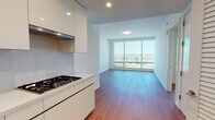 This contemporary high-rise residence offers a clean, modern aesthetic defined by crisp white walls, warm hardwood flooring, and an open, airy layout throughout. The living area is generous and light-filled, anchored by large floor-to-ceiling windows that frame striking panoramic city and mountain views, flooding the space with natural light.

The kitchen is thoughtfully designed with a streamlined, handleless cabinetry system finished in high-gloss white, complemented by light stone countertops and a subtle marble-effect backsplash. It is equipped with stainless steel built-in ovens, a gas cooktop, an integrated sink, and an overhead range hood — providing a fully functional and visually cohesive culinary space.

The bedroom is well-proportioned and calm, also benefiting from large windows with city views, and connects seamlessly to the rest of the unit via clean white-paneled doors. A dedicated hallway provides separation between the living and private areas.

The bathroom is a standout feature, clad entirely in elegant marble-effect tile from floor to ceiling. It includes a wall-hung toilet, a deep soaking tub, and a separate shower enclosure, creating a refined, spa-like atmosphere. Recessed lighting throughout the unit enhances the polished, contemporary ambiance of this well-appointed high-rise residence.
