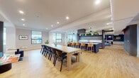 This thoughtfully designed single-floor amenity space offers a diverse range of functional zones suited for both socializing and focused work. The open-plan layout is anchored by a striking communal kitchen, which features dual islands with white countertops, slate-blue cabinetry, open shelving adorned with lush greenery, a statement range hood, and pendant lighting — all finished with light hardwood flooring that runs throughout the entire space.

Adjacent to the kitchen, a large communal dining table with ample seating provides a natural gathering hub, while a bar-height island with warm wood stools offers additional casual seating. The space flows into multiple lounge areas, including a bright, window-lined retreat with panoramic city views, furnished with a generous blue sectional sofa and accent chairs, as well as a separate seating nook with bold green pod chairs and a teal area rug.

A dedicated media lounge offers wall-mounted display screens and a circular arrangement of teal upholstered chairs, ideal for informal presentations or group discussions. For focused individual work, a row of private study alcoves features built-in desks with padded navy acoustic panels, overhead task lighting, and personal seating. Recessed lighting throughout and large windows ensure the space remains bright and inviting.