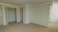 This single-floor space presents a straightforward and functional layout. The bedroom is finished with wall-to-wall carpeting in a warm beige tone, providing a soft underfoot feel throughout the room. The walls are painted in a neutral off-white, creating a clean and versatile backdrop suitable for a variety of furnishing styles. A textured popcorn ceiling adds a classic touch overhead.

A built-in closet is positioned along one wall, featuring an open rod system that offers accessible hanging storage. Adjacent to the closet, a doorway leads to a secondary space, likely a hallway or additional room, indicating a connected and practical floor plan. The room's corner detailing includes a subtle architectural ledge near the ceiling, adding a mild structural element to the otherwise simple design.

Electrical outlets are positioned conveniently along the baseboards, and the overall condition of the space appears clean and ready for occupancy. The neutral finishes and carpeted flooring make this a versatile space adaptable to a range of personal styles and functional needs.
