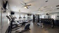 Located in Baton Rouge, Louisiana, this fitness center offers a comprehensive workout environment designed to accommodate a range of exercise routines. The space is housed on a single floor and is organized to maximize functionality while maintaining an open, comfortable atmosphere.

The main workout area features a row of treadmills and elliptical trainers positioned along windowed walls, allowing natural light to filter in through slatted blinds. Multiple ceiling fans are distributed throughout the space, promoting consistent air circulation during workouts. Recessed lighting and wall-mounted televisions add convenience and ambiance.

A dedicated strength training zone includes a multi-station cable machine and a well-stocked dumbbell rack with a full range of free weights, reflected in floor-to-ceiling mirrors that visually expand the space and assist with form monitoring. A flat weight bench accompanies the free weight area for versatile training options.

The entry area includes a water fountain, hand sanitizer dispensers, and posted facility guidelines, indicating this is a 24-hour fitness center. Dark rubber flooring spans the entire space, providing durability and traction. Overall, the fitness center delivers a practical, fully equipped environment suitable for both cardiovascular and strength training workouts.