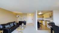 This single-floor residence offers a thoughtfully designed open-concept layout that seamlessly connects the main living areas. The living room is carpeted and comfortably sized, providing a welcoming space for relaxation and entertaining. It is furnished with a leather sofa, a gold-accented coffee table, and complementary accent pieces, all set against warm beige-toned walls that contribute to a cozy ambiance.

A partial dividing wall separates the living room from the kitchen, maintaining an open feel while defining each functional zone. The kitchen features light wood cabinetry, dark countertops, and tile flooring, offering a practical and attractive workspace. A breakfast bar with bar stools provides additional seating and creates a natural gathering point between the kitchen and living area. White appliances, including a dishwasher, are integrated into the cabinetry, and under-cabinet lighting adds brightness to the workspace.

A hallway extends from the living area toward the entry door, providing a clear and accessible path through the home. The overall design balances functionality with a clean, neutral aesthetic, making the space versatile and easy to personalize. The layout is ideal for those seeking an efficient, connected living environment on a single level.