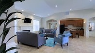 This single-floor space combines a comfortable lounge area with a functional kitchenette, creating an open and inviting environment suited for gatherings or relaxation. The lounge is anchored by a white fireplace along one wall, accompanied by a wall-mounted flat-screen television. Gray upholstered sofas and a plush blue accent chair are arranged around a dark coffee table atop a teal area rug, adding a pop of color to the neutral palette. Large windows allow natural light to fill the room, complementing the recessed lighting overhead.

A graceful arched passageway connects the lounge to an adjacent kitchenette area, which features warm wood cabinetry, dark granite or stone countertops, a black refrigerator, and additional seating along a counter-height bar. The arched entryways and vaulted ceiling add architectural interest, giving the space a sense of openness. Light-toned tile flooring flows throughout, tying the areas together cohesively. The overall ambiance is clean, functional, and comfortable, making it well-suited as a shared or communal space.