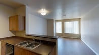 This single-floor space showcases an open-concept layout that seamlessly connects the kitchen and living area. The kitchen features a double stainless steel sink set into a dark laminate countertop, complemented by warm wood cabinetry. A raised breakfast bar divides the kitchen from the adjoining living space, creating a natural boundary while maintaining an open, airy feel. The living area is finished with hardwood-style flooring that runs throughout, adding warmth to the neutral color palette of beige and white walls. Natural light floods the space through a large window fitted with vertical blinds, brightening the room and providing a welcoming ambiance. The textured ceiling adds subtle dimension overhead, while flush-mount ceiling lighting ensures adequate illumination. A secondary window near an entry door further enhances the brightness of the living area. The layout is practical and functional, with clear sightlines from the kitchen to the main living space, making it well-suited for both everyday living and entertaining. The overall design is clean and straightforward, emphasizing comfort and usability.