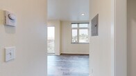 Located in Seattle's Beacon Hill neighborhood, Unit D at 2801 S Hanford Street is a thoughtfully designed, compact single-floor unit that maximizes functionality within its modest footprint. The open layout is unified by light-toned wood-look flooring throughout, contributing to a clean, cohesive aesthetic.

The main living area benefits from multiple windows, including a large multi-pane window and an adjacent balcony door, allowing generous natural light and offering views of the surrounding neighborhood. A recessed ceiling light and an electrical panel are also present in this space.

The kitchen runs along one wall and features white shaker-style cabinetry both above and below a gray countertop, a stainless steel single-basin sink, a smooth-top cooktop, an under-cabinet range hood, and stainless steel appliances including a refrigerator and microwave. The stainless steel backsplash adds a modern, practical touch.

The hallway connects the living area to the bathroom and includes a wall-mounted utility panel. The bathroom is efficiently arranged with wood-look flooring continuing throughout, a toilet, a wall-mounted medicine cabinet with mirror, a grab bar, and a window providing natural ventilation and light. The overall ambiance is clean, bright, and functional.