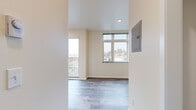 Located in Seattle's Beacon Hill neighborhood, Unit D at 2801 S Hanford Street is a thoughtfully designed, compact single-floor unit that maximizes functionality within its modest footprint. The open layout is unified by light-toned wood-look flooring throughout, contributing to a clean, cohesive aesthetic.

The main living area benefits from multiple windows, including a large multi-pane window and an adjacent balcony door, allowing generous natural light and offering views of the surrounding neighborhood. A recessed ceiling light and an electrical panel are also present in this space.

The kitchen runs along one wall and features white shaker-style cabinetry both above and below a gray countertop, a stainless steel single-basin sink, a smooth-top cooktop, an under-cabinet range hood, and stainless steel appliances including a refrigerator and microwave. The stainless steel backsplash adds a modern, practical touch.

The hallway connects the living area to the bathroom and includes a wall-mounted utility panel. The bathroom is efficiently arranged with wood-look flooring continuing throughout, a toilet, a wall-mounted medicine cabinet with mirror, a grab bar, and a window providing natural ventilation and light. The overall ambiance is clean, bright, and functional.