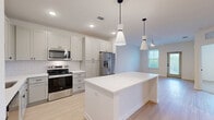 Located at 100 Wilton Cir S in Palm Coast, Florida, this one-bedroom, one-bathroom residence offers a thoughtfully designed single-floor layout with a clean, modern aesthetic throughout. The open-concept living area seamlessly connects the kitchen, dining, and living spaces, all finished with light wood-look flooring that enhances the bright, airy feel of the space. The kitchen is a standout feature, boasting gray shaker-style cabinetry, a white subway tile backsplash, stainless steel appliances including a French door refrigerator, electric range, and microwave, as well as a central island with a white quartz countertop and stylish pendant lighting overhead. The living area offers access to an outdoor balcony and benefits from natural light streaming through multiple windows fitted with blinds, complemented by a ceiling fan. The bedroom is generously sized, featuring plush carpeting, a ceiling fan, and large windows with outdoor views. Three closets provide ample storage throughout the unit. The bathroom is finished with light wood flooring, white cabinetry, a white countertop vanity with matte black hardware, a large mirror, and a glass-enclosed walk-in shower. In-unit laundry adds further convenience to this well-appointed residence.