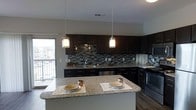This single-floor space showcases a thoughtfully designed kitchen as its centerpiece. Dark espresso cabinetry lines the walls from base to upper level, providing generous storage while creating a bold, contemporary aesthetic. The cabinetry is complemented by a striking mosaic tile backsplash in tones of grey, black, and silver, adding texture and visual interest throughout the cooking area.

The countertops are finished in speckled granite, offering both durability and elegance. A large kitchen island with matching granite extends the prep and dining surface, comfortably accommodating seating for two, as evidenced by the place settings arranged on either side. Two pendant lights with frosted glass shades hang above the island, casting warm, focused illumination that enhances the inviting atmosphere.

Stainless steel appliances, including a dishwasher and range, integrate seamlessly with the dark cabinetry for a cohesive, modern look. A window near the sink and a glass door to what appears to be a balcony or patio allow natural light to filter in, brightening the space. Rich hardwood-style flooring anchors the design, tying together the dark and neutral tones throughout.