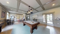 Located at 1001 Bear Island Rd in Summerville, South Carolina, this single-story community clubhouse is thoughtfully designed for gathering, recreation, and relaxation. The centerpiece of the space is a generously sized game room anchored by a solid wood pool table positioned beneath a striking set of industrial-style pendant lights. The room's elegant coffered ceiling adds architectural depth and a refined character throughout. A shuffleboard table lines one wall, offering additional entertainment options for residents and guests. Natural light pours in through large windows dressed with colorful striped valances, framing views of the surrounding greenery. A built-in wet bar serves as a social hub, featuring a dark-topped island with leather bar stools, white subway tile backsplash, a sink, and a wall-mounted flat-screen television — perfect for casual gatherings or community events. An arched chalkboard events board keeps residents informed of upcoming activities. Adjacent to the main game room, a cozy lounge and workspace area provides a more intimate setting with a round café table, vibrant accent chairs, a sofa, and a workstation. Wood-look flooring flows throughout, complemented by wainscoting and crown molding that lend a polished, cohesive aesthetic to the entire space.