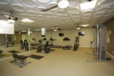 This fitness facility is thoughtfully designed to accommodate a wide range of workout needs within a single-level layout. The space is immediately distinguished by its decorative embossed tile ceiling, which adds a refined aesthetic touch while ceiling fans provide consistent air circulation throughout. Neutral-toned walls and carpeted flooring in the main exercise area create a clean, functional environment.

The cardio zone features multiple Life Fitness treadmills and an elliptical trainer, complemented by a stationary recumbent bike, all positioned for comfortable use. A wall-mounted flat-screen television serves the cardio area. The free weight section includes a well-stocked dumbbell rack, exercise bench, and stability balls, catering to strength training routines.

A cable resistance machine occupies its own dedicated area, and exercise mats are available for floor-based workouts. Floor-to-ceiling mirrors line one wall, visually expanding the space and allowing users to monitor their form. An adjacent area with hardwood flooring, a high-top table, and seating offers a relaxed lounge or stretching zone. Natural light filters in through windows and a glass-paneled entry door, brightening the overall atmosphere.