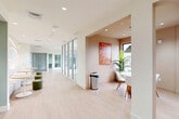 Located at 3424 S University Dr in Davie, Florida, this single-floor commercial space offers a thoughtfully designed interior that balances contemporary aesthetics with functional versatility. Light hardwood flooring runs throughout, lending a clean and cohesive feel to the entire layout.

The hallway serves as the central spine of the space, lined with sleek floor-to-ceiling glass partitions and black metal framing that define private rooms while maintaining an open, transparent atmosphere. Recessed lighting overhead ensures consistent illumination along the full corridor length.

The living and lounge area is a standout feature, highlighted by a dramatic sculptural wall with flowing, backlit wave reliefs that create an inviting and visually striking focal point. White upholstered banquette seating curves along the wall, complemented by round light wood tables and green fabric ottomans, offering a comfortable and stylish gathering area.

The dining room is a warm, intimate nook bathed in natural light from a large window framing outdoor views of the surrounding landscape. A round wood dining table, modern white chairs, a sage green pendant light, and vibrant wall art complete the space, adding personality and color.

At the far end, a spacious open room with floor-to-ceiling glass doors leads to an outdoor balcony, offering panoramic views and abundant natural light.