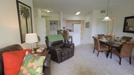 This single-floor residence offers a well-designed open-concept floor plan that integrates the living, dining, and kitchen areas into one cohesive, comfortable space. The living area is carpeted and generously proportioned, providing ample room for relaxed seating arrangements, as evidenced by the plush leather recliner and sofa accented with tropical-patterned cushions that lend a warm, casual ambiance to the space.

Adjacent to the living area, the dining space features wicker-style chairs gathered around a round table, complementing the natural, relaxed aesthetic carried throughout the unit. The kitchen is efficiently designed with light wood cabinetry, a white refrigerator, and a breakfast bar with bar-height seating, making it both functional and sociable. Recessed overhead lighting illuminates the kitchen, while flush-mount fixtures provide consistent lighting throughout the main living areas.

A standout feature of this residence is the dedicated laundry area, visible through a doorway off the main living space, offering the convenience of an in-unit washer and dryer. The consistent use of neutral tones on the walls and carpeted flooring ties the entire space together, creating a cohesive and inviting atmosphere ideal for comfortable everyday living.