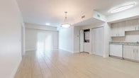 This single-floor unit offers a clean, modern aesthetic defined by light-toned tile flooring, crisp white walls, and abundant natural light throughout. The open-concept living and dining area is spacious and airy, anchored by an elegant pendant light fixture and complemented by vertical blinds that allow soft, diffused light to filter in from a sliding glass door. A stacked washer and dryer unit is conveniently tucked behind a louvered closet door within the main living space, adding practical in-unit laundry access. The kitchen is thoughtfully designed with light wood-tone cabinetry, granite-style countertops, and white appliances including a refrigerator, electric range, and microwave. A dedicated pantry with open shelving provides generous storage. The bedroom is generously sized, featuring two windows dressed with vertical blinds that flood the room with natural light. The bathroom maintains the unit's clean, bright palette with light tile flooring and a grab bar installed for added accessibility. Throughout the unit, the consistent use of light materials and neutral tones creates a cohesive, welcoming atmosphere that feels open and easy to personalize.