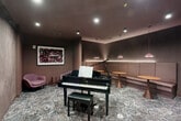 Located in Everett, MA, this thoughtfully designed music room offers an intimate yet refined setting for performance, listening, and socializing. The centerpiece of the space is a polished black Yamaha grand piano, positioned beneath recessed ceiling lights that illuminate the room with a warm, focused glow. The flooring throughout is a striking veined marble in cool gray and brown tones, lending an air of sophistication to the entire space.

The room is enveloped in a muted mauve-and-taupe color palette, creating a cohesive and calming ambiance. A padded banquette runs along one wall, accompanied by round amber-toned pedestal tables and suspended dome pendant lights in a complementary rose hue, forming a comfortable lounge area for guests. A plush pink accent chair is tucked into a corner beside a bold abstract artwork, while a circular sculptural wall piece adds a dramatic focal point near the piano.

Along one wall, dark cabinetry with brushed gold hardware frames a built-in bar area, complete with under-counter refrigeration and a decorative backsplash, making this room equally suited for intimate social events. The overall design balances artistic expression with functional comfort, resulting in a space that is both visually striking and acoustically purposeful.