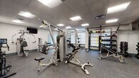 Located at 2 Broadway in Lynnfield, Massachusetts, this fitness center offers a well-equipped, single-floor workout facility designed to accommodate a wide range of exercise routines. The space features a drop ceiling with recessed panel lighting that provides bright, even illumination throughout, while durable rubber flooring offers comfort and safety underfoot.

The strength training area is outfitted with a multi-station cable machine, a power rack, a dedicated weight bench, and a well-stocked dumbbell rack with varying weights. Medicine balls and resistance tools are neatly organized on wall-mounted storage racks, keeping the space tidy and accessible.

The cardio section includes multiple Cybex treadmills and a stationary spin bike equipped with a digital display console, catering to users of all fitness levels. A wall-mounted flat-screen television is positioned above a large floor-to-ceiling mirror, which expands the perceived size of the room and allows users to monitor their form during workouts.

The facility also features an upper-body cable and arm workout machine, adding further versatility to the training options available. The neutral color palette of white and gray walls, combined with the reflective mirrors, contributes to a clean, professional atmosphere. A private door provides access to an adjacent room, rounding out this fully functional fitness environment.