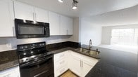 This single-floor space showcases a well-appointed kitchen designed for both functionality and style. The kitchen features crisp white shaker-style cabinetry with sleek bar-pull hardware, providing ample upper and lower storage. Dark granite countertops offer a striking contrast against the white cabinets, lending the space a contemporary aesthetic. Black appliances, including an electric range and over-the-range microwave, complement the bold countertop tones. A double stainless steel sink with a modern pull-down faucet is conveniently positioned at the corner, maximizing workspace efficiency. Light hardwood-style flooring grounds the kitchen, transitioning seamlessly into the adjacent living area, which benefits from neutral-toned carpet and an open, airy feel. Recessed track lighting illuminates the kitchen workspace effectively. The open-concept layout connects the kitchen directly to the living space, enhancing the overall sense of flow and making the area well-suited for everyday living and entertaining. The neutral color palette throughout creates a clean, inviting ambiance.