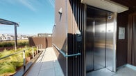 This rooftop terrace is a well-appointed outdoor space defined by a cohesive contemporary aesthetic. The exterior façade and privacy screening are clad in dark corrugated metal panels, lending the space a bold, industrial character that complements its urban setting. Large-format light stone pavers cover the terrace floor throughout, providing a clean and durable surface underfoot.

A standout feature of this space is its open-air seating area, furnished with wooden lounge chairs positioned to take full advantage of sweeping panoramic views of the surrounding cityscape, including rooftops, hills, and the broader urban skyline. Raised planter beds, finished in a warm corten-style material, are filled with ornamental grasses and low-maintenance succulents, introducing natural greenery that softens the industrial palette.

Perimeter safety railings constructed from galvanized steel mesh framing provide unobstructed sightlines across the city while maintaining a secure boundary. The terrace is served by a stainless steel elevator, offering convenient access to and from the building below. A hand sanitizer station is positioned near the elevator lobby, reflecting a commitment to hygiene and accessibility.

An overhead pergola-style canopy structure provides partial shade near the elevator entry point. The space benefits from abundant natural light and open sky, making it an inviting outdoor retreat above the city.