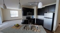 This single-floor space showcases a thoughtfully designed open-concept layout where the kitchen serves as a central focal point. The kitchen features striking dark cabinetry that contrasts elegantly with light granite countertops, creating a bold, contemporary aesthetic. A double-basin stainless steel sink is prominently positioned on the kitchen island, which also provides additional prep space and casual seating potential.

The appliance suite includes a stainless steel electric range, an over-the-range microwave, and a full-size refrigerator, all integrated seamlessly into the dark cabinetry design. A classic white subway tile backsplash adds a clean, polished detail to the cooking area. Track lighting on the ceiling, complemented by two pendant lights hanging above the island, ensures the kitchen is well-lit and functional.

The kitchen opens naturally into a spacious living area, which benefits from natural light streaming through a window. The light-toned walls and flooring of the adjacent living space provide a pleasing contrast to the kitchen's darker palette, giving the overall space a balanced and inviting ambiance.