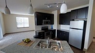 This single-floor space showcases a thoughtfully designed open-concept layout where the kitchen serves as a central focal point. The kitchen features striking dark cabinetry that contrasts elegantly with light granite countertops, creating a bold, contemporary aesthetic. A double-basin stainless steel sink is prominently positioned on the kitchen island, which also provides additional prep space and casual seating potential.

The appliance suite includes a stainless steel electric range, an over-the-range microwave, and a full-size refrigerator, all integrated seamlessly into the dark cabinetry design. A classic white subway tile backsplash adds a clean, polished detail to the cooking area. Track lighting on the ceiling, complemented by two pendant lights hanging above the island, ensures the kitchen is well-lit and functional.

The kitchen opens naturally into a spacious living area, which benefits from natural light streaming through a window. The light-toned walls and flooring of the adjacent living space provide a pleasing contrast to the kitchen's darker palette, giving the overall space a balanced and inviting ambiance.