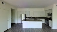 This single-floor space showcases a thoughtfully designed, open-concept kitchen that serves as the focal point of the layout. The kitchen features an expansive run of white shaker-style cabinetry that extends from floor to ceiling, providing generous storage throughout. Dark, matte countertops offer a striking contrast against the bright white cabinetry, lending the space a contemporary and sophisticated aesthetic. A large central island with a matching dark countertop anchors the kitchen, offering additional workspace and the potential for casual seating. Stainless steel appliances, including a full-size refrigerator, are seamlessly integrated into the cabinetry. A dedicated sink with a modern faucet is positioned along the back wall, flanked by additional counter space. Recessed lighting is distributed evenly across the white ceiling, ensuring the space is well-lit throughout. Dark hardwood flooring runs continuously across the open area, adding warmth and depth while complementing the overall color palette. The open layout promotes a natural flow between kitchen and living areas, making the space both functional and inviting.