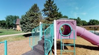 This outdoor space centers on a well-equipped playground area designed for children, situated on a generous wood chip safety surface that surrounds the play equipment. The main play structure is finished in purple and pink tones and features a central play pod with a circular tunnel entrance, a curved slide extending from one side, and a vertical ladder providing an additional access point. A set of wide metal stairs with teal-colored railings leads up to a platform connected to the main pod, offering an accessible entry route. The teal railings and metal accents throughout the structure provide a consistent, vibrant color scheme against the neutral wood chip ground cover.

The playground is set within a well-maintained green lawn, with mature evergreen and deciduous trees forming a natural backdrop that provides shade and a pleasant outdoor atmosphere. A brick building with a gambrel-style roof and brown shingles is visible nearby, indicating the playground is associated with a residential or community property. The grounds are neatly kept, with paved pathways running alongside the grassy areas. The open sky and surrounding tree canopy create a bright, welcoming environment well-suited for outdoor recreation and play.