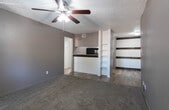 Located at 4900 E University Blvd in Odessa, Texas, this single-floor unit offers a functional and cohesive layout finished in a consistent neutral gray palette throughout. The living area features plush carpeting underfoot and is anchored by a ceiling fan with integrated lighting. A sliding glass door floods the space with natural light and provides direct access to an outdoor area, offering views of surrounding greenery. The transition between the living space and kitchen is defined by a half-wall pass-through counter with a dark wood-tone top, complemented by built-in open shelving and rustic wood accent rails — a distinctive design feature that adds character to the space. The kitchen is equipped with black appliances, including a refrigerator and range, set against light wood-look flooring and white cabinetry with granite-style countertops. A hallway connects the main living areas to the bedrooms, each of which is carpeted and includes a ceiling fan with lighting and windows fitted with vertical blinds that allow for natural light. The bathroom is well-appointed with wood-look flooring, a single-sink vanity with granite-style countertop and a framed mirror, along with a full tub-shower combination with white tile surround. Textured ceilings and gray walls tie the entire unit together with a modern, understated aesthetic.