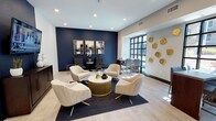 This elegantly designed single-floor space functions as a upscale communal lounge and meeting area, blending contemporary aesthetics with functional versatility. The layout is thoughtfully divided into multiple zones, each serving a distinct purpose while maintaining a cohesive design language throughout.

The central lounge area anchors the space with a cluster of plush, cream-upholstered swivel chairs arranged around a gold-ribbed circular coffee table with a marble top, all set atop a navy patterned area rug over light wood-style flooring. A dramatic navy accent wall provides a rich backdrop, adorned with a triptych of abstract art in gold-toned frames, complemented by tufted leather accent chairs beneath.

A secondary seating nook features whitewashed exposed brick walls, a wall-mounted television, and additional lounge seating, adding textural contrast. Adjacent to the main lounge, a high-top bar-style table with sleek gray chairs sits beneath a large, grid-paned window that floods the area with natural light and exterior views. Sculptural gold disc wall art nearby adds an artistic focal point.

Glass-paneled double doors lead to a private conference or meeting room, enhancing the space's functionality. Recessed lighting throughout ensures a warm, polished ambiance.