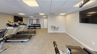 This fitness center offers a comprehensive, single-floor workout facility designed to accommodate a wide range of exercise routines. The space is bright and open, with large windows along one wall that allow ample natural light to fill the room and provide views of the surrounding outdoor scenery, creating an inviting atmosphere for exercise.

The cardio area is well-stocked with multiple treadmills, elliptical machines, and stationary bikes arranged in rows, all oriented toward wall-mounted flat-screen televisions for an engaging workout experience. A multi-station cable strength machine is also present, offering versatile resistance training options.

The free weights section features a full rack of dumbbells in varying sizes, accompanied by an adjustable workout bench, providing a dedicated space for strength training. A water fountain station with a dual-height design is conveniently located within the facility, ensuring easy hydration access for all users.

The flooring throughout consists of durable, light-toned tile and carpet tile, well-suited for a high-traffic fitness environment. The drop ceiling features recessed panel lighting that ensures consistent, even illumination throughout the space. Neutral gray and beige tones on the walls contribute to a clean, modern aesthetic. A dedicated entrance door and a trash receptacle complete the practical amenities of this thoughtfully appointed fitness center.