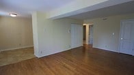 This single-floor residence offers a clean, functional layout designed for comfortable everyday living. The main living area is highlighted by rich, warm-toned hardwood flooring that extends throughout the space, creating a cohesive and inviting atmosphere. Neutral, light beige walls provide a versatile backdrop that complements a variety of interior styles and furnishings.

Structural elements such as a central dividing wall with a white paneled door help define distinct zones within the open layout, offering a sense of separation between the living area and the adjoining rooms beyond. The white trim and baseboards throughout provide a crisp contrast against the warm flooring and soft wall tones, contributing to the overall tidiness of the space.

Doorways visible from the main area lead to additional rooms, suggesting a well-connected floor plan that flows naturally from one space to the next. The ceiling is smooth and standard height, keeping the interior feel grounded and practical. Overhead ventilation is present, indicating a climate-controlled environment for year-round comfort.

Overall, this residence presents a straightforward, well-maintained interior with classic finishes, making it a solid and adaptable space suitable for a range of occupants and furnishing styles.