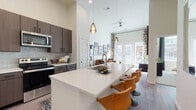 This contemporary single-floor residence showcases a thoughtfully designed open-concept layout, blending modern finishes with functional living spaces throughout. The kitchen is a standout feature, offering rich dark wood cabinetry with matte black hardware, sleek white quartz countertops, a full suite of stainless steel appliances including a refrigerator, electric range, microwave, and dishwasher, and a tile backsplash in a soft blue-gray tone. A generously sized center island with an integrated sink provides ample prep space and doubles as a casual dining area, complemented by pendant lighting overhead. The open living area benefits from soaring ceilings and an abundance of natural light streaming through full-height French doors that lead to an outdoor area. The bedroom is a comfortable retreat, featuring large windows that frame views of the surrounding community and offer excellent natural light. The bathroom includes a tub-shower combination with floor-to-ceiling tile surround, wood-toned cabinetry, and hardwood-style flooring. A dedicated laundry closet houses a stacked Whirlpool washer and dryer for added convenience. Hardwood-style flooring flows throughout the main living areas, tying together the modern, cohesive aesthetic of this well-appointed residence.