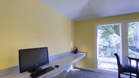 This single-floor space is designed with functionality and simplicity in mind. The room features warm yellow walls that lend a bright, energetic atmosphere, complemented by a white textured ceiling that adds a subtle visual interest overhead. A sleek, wall-mounted built-in desk runs along one side of the room, providing an efficient and streamlined workspace. The desk is equipped with a desktop computer, keyboard, and mouse, making it well-suited for productivity. A small decorative blue vase adds a touch of personality to the otherwise minimal setup. Patterned carpet tiles cover the floor, contributing a professional and practical feel underfoot. Natural light filters generously into the space through a large window positioned along one wall, offering a pleasant view of lush outdoor greenery. The window not only brightens the interior but also creates a refreshing connection to the outdoors. The overall layout is compact yet thoughtfully arranged, maximizing the use of available space while maintaining an open and uncluttered feel. This space is well-suited as a private office or dedicated study area.