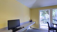 This single-floor space is designed with functionality and simplicity in mind. The room features warm yellow walls that lend a bright, energetic atmosphere, complemented by a white textured ceiling that adds a subtle visual interest overhead. A sleek, wall-mounted built-in desk runs along one side of the room, providing an efficient and streamlined workspace. The desk is equipped with a desktop computer, keyboard, and mouse, making it well-suited for productivity. A small decorative blue vase adds a touch of personality to the otherwise minimal setup. Patterned carpet tiles cover the floor, contributing a professional and practical feel underfoot. Natural light filters generously into the space through a large window positioned along one wall, offering a pleasant view of lush outdoor greenery. The window not only brightens the interior but also creates a refreshing connection to the outdoors. The overall layout is compact yet thoughtfully arranged, maximizing the use of available space while maintaining an open and uncluttered feel. This space is well-suited as a private office or dedicated study area.