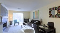 This single-floor residence offers a thoughtfully designed living space that balances comfort and modern style. The main living area serves as the centerpiece of the home, featuring plush wall-to-wall carpeting that adds warmth underfoot. Large sliding glass doors flood the room with natural light, creating an airy and inviting atmosphere while providing a seamless connection to the outdoors. Vertical blinds offer flexible light control throughout the day. The living area is well-proportioned, comfortably accommodating a tufted dark sofa, accent seating, and a sleek coffee table arranged around a soft area rug. A bold, colorful abstract artwork anchors one of the neutral-toned walls, adding a vibrant focal point to the otherwise understated palette of soft whites and warm beiges. A dedicated workspace or dining area is visible near the entrance, featuring a dark desk and chair, highlighting the functional versatility of the layout. The overall ambiance is calm and contemporary, with clean lines, minimal clutter, and an emphasis on natural light — making the space feel open, organized, and move-in ready.