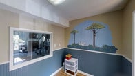 This single-floor space offers a uniquely creative environment highlighted by a standout safari-themed room. The room's most striking feature is a vibrant, hand-painted wall mural depicting a lush savanna scene, complete with tall trees, leafy green foliage, a zebra grazing among the bushes, and a monkey swinging from the branches. The mural spans a large portion of the wall, adding an immersive, artistic character to the space.

The room features a two-tone wall treatment, with the lower half finished in a classic beadboard-style blue paneling and the upper half painted in a warm tan tone, creating a structured and visually appealing contrast. The flooring is warm hardwood, contributing to an inviting atmosphere. A textured ceiling adds additional depth to the overall design.

An interior window connects this room to an adjacent fitness area, which is equipped with a multi-station weight machine, allowing visibility between the two spaces and creating a sense of openness. The fitness area is well-appointed for strength training and exercise.

Together, the two areas reflect a thoughtfully designed space that balances playful creativity with practical functionality, making it suitable for both leisure and active use.