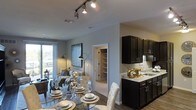 This single-floor residence offers an open-concept layout that seamlessly connects the kitchen, dining area, and living room into one cohesive living space. The kitchen is outfitted with dark espresso cabinetry, light granite countertops, and a stainless steel sink, creating a striking contrast that anchors the space. Track lighting runs along the ceiling, providing focused illumination throughout the open area and complementing the contemporary aesthetic.

The dining area features a glass-top table with upholstered white chairs, positioned to take full advantage of the flowing layout. Adjacent to it, the living room is warmly lit by a combination of floor lamps and pendant lighting, giving the area an inviting ambiance. A large sliding glass door or window leads to a private balcony, allowing natural light to filter into the space.

A doorway visible from the main living area leads to what appears to be a walk-in closet or storage space with built-in shelving, adding practical functionality. The neutral gray walls and wood-style flooring unify the space, contributing to a clean, modern atmosphere throughout.