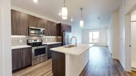 This single-floor residence at 7323 W 38th Ave in Wheat Ridge, Colorado, showcases a contemporary design with a clean, open-concept layout. The kitchen serves as a central highlight, featuring rich dark-wood cabinetry extending to the ceiling, a subway tile backsplash, and a full suite of stainless steel appliances including a French door refrigerator, electric range, and microwave. A spacious island with a white quartz countertop and undermount sink anchors the space, illuminated by sleek pendant lighting overhead. Wood-look plank flooring flows seamlessly from the kitchen into the open living area, which benefits from natural light through large windows with outdoor views and ample room for a variety of furniture arrangements. The bathroom is well-appointed with a dual-sink vanity featuring dark cabinetry and light countertops, a large mirror, and a walk-in shower finished with classic white subway tile. Bedroom spaces offer a mix of wood-look and soft carpet flooring, with windows providing natural light and exterior views. Throughout the unit, crisp white walls and modern finishes create a bright, cohesive ambiance well-suited to contemporary living in the Wheat Ridge area.