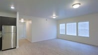 This single-floor space is designed with a clean, minimalist aesthetic that emphasizes openness and functionality. The main living area features light hardwood-style flooring that extends throughout, complementing the crisp white walls and creating a bright, airy atmosphere. Two side-by-side windows dressed with horizontal blinds allow natural light to filter in, illuminating the room evenly and enhancing the sense of spaciousness.

Flush-mount ceiling light fixtures are positioned throughout, ensuring consistent artificial lighting across the space. The smooth, neutral-toned ceilings add to the overall clean appearance of the interior. A centrally positioned partial wall divides the main living area from an adjoining hallway or room, providing a subtle architectural separation while maintaining an open flow between spaces.

The hallway visible in the background suggests additional rooms beyond the main living area, with continued use of the same light flooring and neutral palette. White baseboards frame the perimeter of the room, adding a polished finishing detail. Overall, the space presents a well-lit, move-in-ready interior with a cohesive, understated design throughout.
