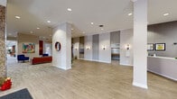 Located at 25 Washington Ln in Wyncote, Pennsylvania, this generously sized single-floor space serves as a welcoming and well-appointed lobby and common area. The entrance features floor-to-ceiling glass panel doors framed in dark metal, flanked by textured stone accent walls that set an elegant tone from the moment of arrival. Recessed lighting throughout maintains a bright, clean atmosphere across all areas.

The reception area is anchored by a sleek, modern white desk with ample workspace, positioned beneath a coffered skylight panel that draws in natural light. Adjacent to the entry, a spacious lounge area features neutral-toned patterned carpeting, built-in stone bench seating with colorful accent cushions, and large windows overlooking lush outdoor greenery. A separate sitting area is furnished with vibrant blue upholstered chairs, warm cognac leather sofas, and a wood slab coffee table, accented by contemporary artwork on the walls.

The hallways are finished with light wood-look luxury vinyl flooring, crown molding, and recessed lighting, providing clean circulation paths throughout. Elevator alcoves are tastefully designed with wall sconces and panel surrounds. Rich wood detailing, including a prominent staircase banister, adds warmth and character. The overall ambiance is polished, modern, and inviting.