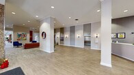 Located at 25 Washington Ln in Wyncote, Pennsylvania, this generously sized single-floor space serves as a welcoming and well-appointed lobby and common area. The entrance features floor-to-ceiling glass panel doors framed in dark metal, flanked by textured stone accent walls that set an elegant tone from the moment of arrival. Recessed lighting throughout maintains a bright, clean atmosphere across all areas.

The reception area is anchored by a sleek, modern white desk with ample workspace, positioned beneath a coffered skylight panel that draws in natural light. Adjacent to the entry, a spacious lounge area features neutral-toned patterned carpeting, built-in stone bench seating with colorful accent cushions, and large windows overlooking lush outdoor greenery. A separate sitting area is furnished with vibrant blue upholstered chairs, warm cognac leather sofas, and a wood slab coffee table, accented by contemporary artwork on the walls.

The hallways are finished with light wood-look luxury vinyl flooring, crown molding, and recessed lighting, providing clean circulation paths throughout. Elevator alcoves are tastefully designed with wall sconces and panel surrounds. Rich wood detailing, including a prominent staircase banister, adds warmth and character. The overall ambiance is polished, modern, and inviting.