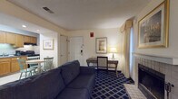 Located at 246 Daniel Webster Highway in Merrimack, New Hampshire, this generously sized hotel suite offers a well-rounded layout designed for both comfort and practicality. The main living area serves as the centerpiece of the suite, furnished with a plush sofa, a pedestal coffee table, and a flat-screen television mounted atop a tall dresser. A standout feature of the living space is the tiled wood-burning fireplace, which adds warmth and character to the room. Framed artwork adorns the cream-colored walls throughout, contributing to a cohesive, welcoming atmosphere. A dedicated work area includes a wooden desk, lamp, and chair — well-suited for business travelers. Two beds provide ample sleeping accommodations, positioned on bold navy blue carpeting with a geometric pattern that runs throughout the suite. A fully equipped kitchen features warm oak cabinetry, black appliances including a full-size refrigerator, stove, microwave, and dishwasher, as well as a dining table with seating for two. The bathroom offers a granite-topped vanity, large mirror, and wall-mounted hair dryer. Natural light filters through draped windows with valances, enhancing the suite's comfortable, residential feel.