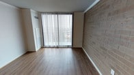 This single-floor space showcases a clean, functional design highlighted by several distinctive architectural elements. The main living area features smooth wood-look laminate flooring that runs throughout, lending warmth and continuity to the space. A full-height exposed brick accent wall serves as a striking focal point, adding texture and character to the otherwise neutral interior. Large sliding glass doors, dressed with vertical blinds, flood the room with natural light while providing direct access to an outdoor balcony, offering views of the surrounding area. The white walls and light ceiling create a bright, open atmosphere that complements the natural tones of both the flooring and brick. A wall-mounted HVAC unit and visible ceiling vent indicate a dedicated climate control system built into the unit. The overall ambiance is understated and versatile, offering a neutral canvas well-suited for a variety of furnishing styles. The combination of natural light, textural contrast, and durable finishes makes this a practical yet visually appealing space.