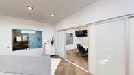 This single-floor office suite is thoughtfully designed to maximize functionality within a compact footprint. The space is divided into two distinct zones, seamlessly connected by sliding frosted-glass partition doors that allow natural light to filter between areas while maintaining privacy when needed.

The open workstation area features herringbone-patterned flooring, white walls, and recessed lighting overhead. A panel-divided desk provides a focused workspace, and a white storage cabinet topped with a printer and a potted snake plant adds practical utility. Interior windows maintain visual connectivity between zones.

The private conference room is a polished, well-appointed meeting space anchored by a light wood rectangular table surrounded by teal velvet chairs with gold-toned bases. A wall-mounted flat-screen display and a striking gold sputnik-style chandelier serve as functional focal points. A painted white brick accent wall adds texture and character, while a wall-mounted mini-split HVAC unit ensures climate comfort. Tall indoor greenery adds a welcoming, organic touch.

Throughout, the design maintains a cohesive, modern aesthetic with an all-white color palette, herringbone flooring, and carefully curated furnishings — creating a professional yet inviting environment well-suited for focused work and collaborative meetings alike.