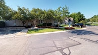 Crooked Creek is a single-story property that makes a strong first impression with its thoughtfully maintained exterior. The building features a neutral-toned siding exterior that blends seamlessly with its surroundings, while the façade is complemented by classic wall-mounted light fixtures that flank a central entrance door. The approach to the building is defined by a clean, paved pathway leading directly to the front entry, flanked by meticulously manicured landscaping. Vibrant seasonal flower beds in rich reds and oranges line the front of the building, adding striking color and curb appeal. Mature deciduous trees are strategically placed across the property, providing natural shade and a sense of established character. The surrounding grounds are kept in pristine condition, with neatly trimmed grass and well-defined planting areas that contribute to an inviting, professional appearance. Ample paved parking is available directly in front of the building, offering convenient access for visitors. The overall ambiance of the property is polished and approachable, projecting a welcoming atmosphere suited for a variety of uses. The single-floor layout ensures easy accessibility throughout, making this a practical and visually appealing space.