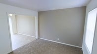 This two-story residence offers a clean, straightforward layout with a neutral color palette throughout. The interiors are finished with warm beige and greige tones on the walls, complemented by white trim and ceilings that lend a bright, cohesive feel to the space. The living area is carpeted in a muted, natural tone, providing a soft underfoot feel and a welcoming atmosphere. An open doorway connects the main living space to an adjacent area finished with tile or hard flooring, offering a practical transition between zones and adding visual contrast to the interior. The walls are smooth and simply detailed, with clean white baseboards framing each room. Electrical outlets are conveniently placed, and ceiling vents suggest a central HVAC system for year-round climate control. The overall design is understated and move-in ready, providing a blank canvas well-suited for personalization. Spanning two floors, the home offers separation between living and private spaces, making it a practical and versatile layout for a range of occupants.
