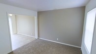 This two-story residence offers a clean, straightforward layout with a neutral color palette throughout. The interiors are finished with warm beige and greige tones on the walls, complemented by white trim and ceilings that lend a bright, cohesive feel to the space. The living area is carpeted in a muted, natural tone, providing a soft underfoot feel and a welcoming atmosphere. An open doorway connects the main living space to an adjacent area finished with tile or hard flooring, offering a practical transition between zones and adding visual contrast to the interior. The walls are smooth and simply detailed, with clean white baseboards framing each room. Electrical outlets are conveniently placed, and ceiling vents suggest a central HVAC system for year-round climate control. The overall design is understated and move-in ready, providing a blank canvas well-suited for personalization. Spanning two floors, the home offers separation between living and private spaces, making it a practical and versatile layout for a range of occupants.